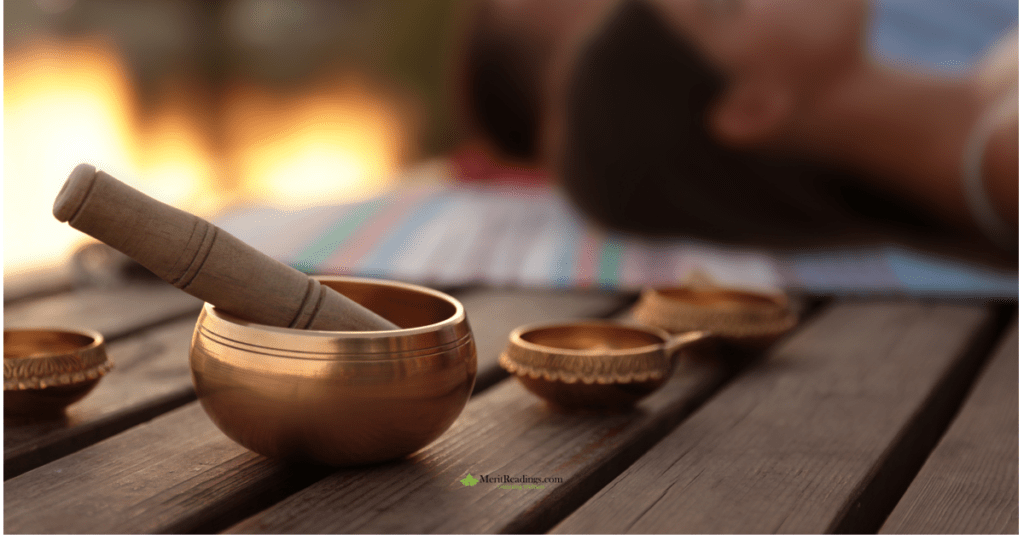  A brass singing bowl with a wooden mallet placed on a wooden surface, with smaller decorative bowls nearby and a blurred background showing a person lying down.